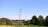 A landscape featuring tall metal electricity pylons extending into the distance, set against a clear blue sky. Lush greenery is present in the foreground, with trees scattered throughout, creating a serene and vast natural scene.