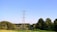 A landscape featuring tall metal electricity pylons extending into the distance, set against a clear blue sky. Lush greenery is present in the foreground, with trees scattered throughout, creating a serene and vast natural scene.