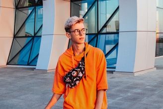 A young person with short blond hair and glasses wears an orange hoodie and a black patterned crossbody bag. They stand in front of a modern building with geometric glass and metal design.