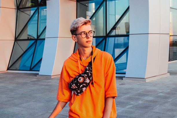 A young person with short blond hair and glasses wears an orange hoodie and a black patterned crossbody bag. They stand in front of a modern building with geometric glass and metal design.