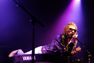 A musician with curly hair and sunglasses is singing into a microphone while lying on top of a Yamaha keyboard. The setting is a stage with dramatic purple and pink lighting, creating a vibrant atmosphere.