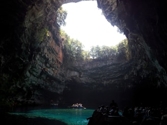 A large underground cave with a circular opening at the top, allowing sunlight to illuminate the scene. The interior features rugged rock walls covered with vegetation. At the bottom, clear turquoise water reflects the light above, and a small group of people can be seen in boats on the water, enhancing the sense of exploration and wonder.