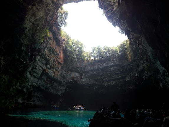 A large underground cave with a circular opening at the top, allowing sunlight to illuminate the scene. The interior features rugged rock walls covered with vegetation. At the bottom, clear turquoise water reflects the light above, and a small group of people can be seen in boats on the water, enhancing the sense of exploration and wonder.