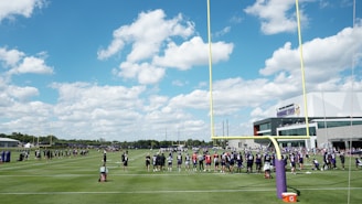 A group of football players, wearing uniforms, are lined up on a green field with a bright blue sky and white clouds in the background. There is a large sports facility building to the right, and the scene is framed by yellow goalposts. Equipment and a water cooler can be seen on the field.