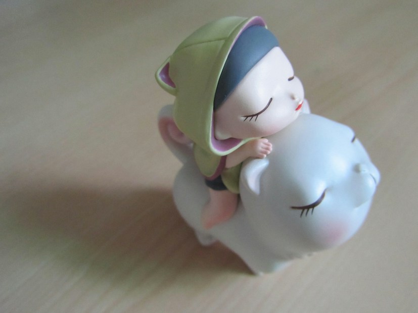 A small figurine depicting a childlike character with closed eyes wearing a green hood, sitting on a large, white, round-bodied animal with a cat-like appearance. Both appear relaxed, suggesting a peaceful or dreamlike scene.