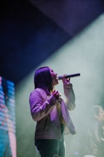 A person is singing passionately on stage, holding a microphone. The spotlight casts a dramatic glow around them, and smoke effects are visible in the background. The atmosphere suggests a live performance with vibrant lighting.