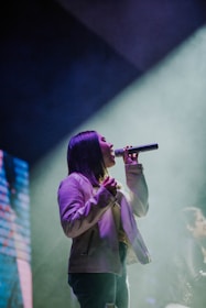 A person is singing passionately on stage, holding a microphone. The spotlight casts a dramatic glow around them, and smoke effects are visible in the background. The atmosphere suggests a live performance with vibrant lighting.