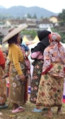 A group of women dressed in traditional, colorful clothing are gathered outdoors, engaging in conversation. One woman wears a wide-brimmed hat, and another holds a child. The background features a hilly landscape and a few structures.
