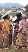 A group of women dressed in traditional, colorful clothing are gathered outdoors, engaging in conversation. One woman wears a wide-brimmed hat, and another holds a child. The background features a hilly landscape and a few structures.