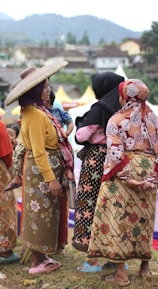 A group of women dressed in traditional, colorful clothing are gathered outdoors, engaging in conversation. One woman wears a wide-brimmed hat, and another holds a child. The background features a hilly landscape and a few structures.