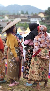 A group of women dressed in traditional, colorful clothing are gathered outdoors, engaging in conversation. One woman wears a wide-brimmed hat, and another holds a child. The background features a hilly landscape and a few structures.