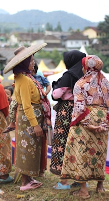 A group of women dressed in traditional, colorful clothing are gathered outdoors, engaging in conversation. One woman wears a wide-brimmed hat, and another holds a child. The background features a hilly landscape and a few structures.