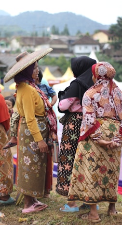 A group of women dressed in traditional, colorful clothing are gathered outdoors, engaging in conversation. One woman wears a wide-brimmed hat, and another holds a child. The background features a hilly landscape and a few structures.