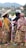 A group of women dressed in traditional, colorful clothing are gathered outdoors, engaging in conversation. One woman wears a wide-brimmed hat, and another holds a child. The background features a hilly landscape and a few structures.