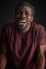 A person with an expressive face, possibly laughing or displaying a strong emotion, wearing a maroon shirt. The lighting is dim with a focus on the face and upper body against a black background.