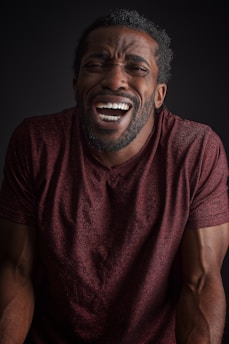 A person with an expressive face, possibly laughing or displaying a strong emotion, wearing a maroon shirt. The lighting is dim with a focus on the face and upper body against a black background.