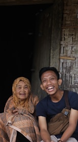 An elderly woman wearing a patterned headscarf and a young man with a bag across his shoulder are sitting together. They both have joyful expressions and are engaging directly with the camera. The background shows a rustic wooden structure.