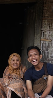 An elderly woman wearing a patterned headscarf and a young man with a bag across his shoulder are sitting together. They both have joyful expressions and are engaging directly with the camera. The background shows a rustic wooden structure.