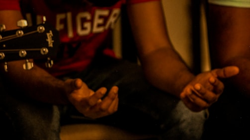 A close-up of hands gesturing with slightly open palms, alongside a part of an acoustic guitar. The scene is warmly lit, focusing on the expressive body language.