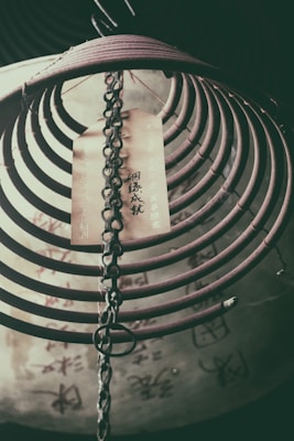 A coiled incense burner hangs with a chain, featuring an attached paper strip with Chinese characters. The background reveals more characters on a blurred surface, suggesting a traditional or cultural setting.