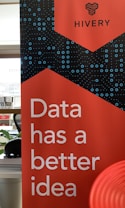 A promotional banner with a bold red background features the text 'Data has a better idea' in large white letters. Above the text, the word 'HIVERY' is printed along with a stylized logo resembling a beehive. The background also includes a pattern of blue circles and dots, giving a digital or technological feel. In the surrounding area, there's a partial view of a desk and some office supplies.