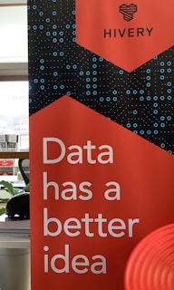 A promotional banner with a bold red background features the text 'Data has a better idea' in large white letters. Above the text, the word 'HIVERY' is printed along with a stylized logo resembling a beehive. The background also includes a pattern of blue circles and dots, giving a digital or technological feel. In the surrounding area, there's a partial view of a desk and some office supplies.