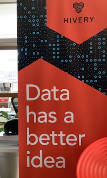 A promotional banner with a bold red background features the text 'Data has a better idea' in large white letters. Above the text, the word 'HIVERY' is printed along with a stylized logo resembling a beehive. The background also includes a pattern of blue circles and dots, giving a digital or technological feel. In the surrounding area, there's a partial view of a desk and some office supplies.