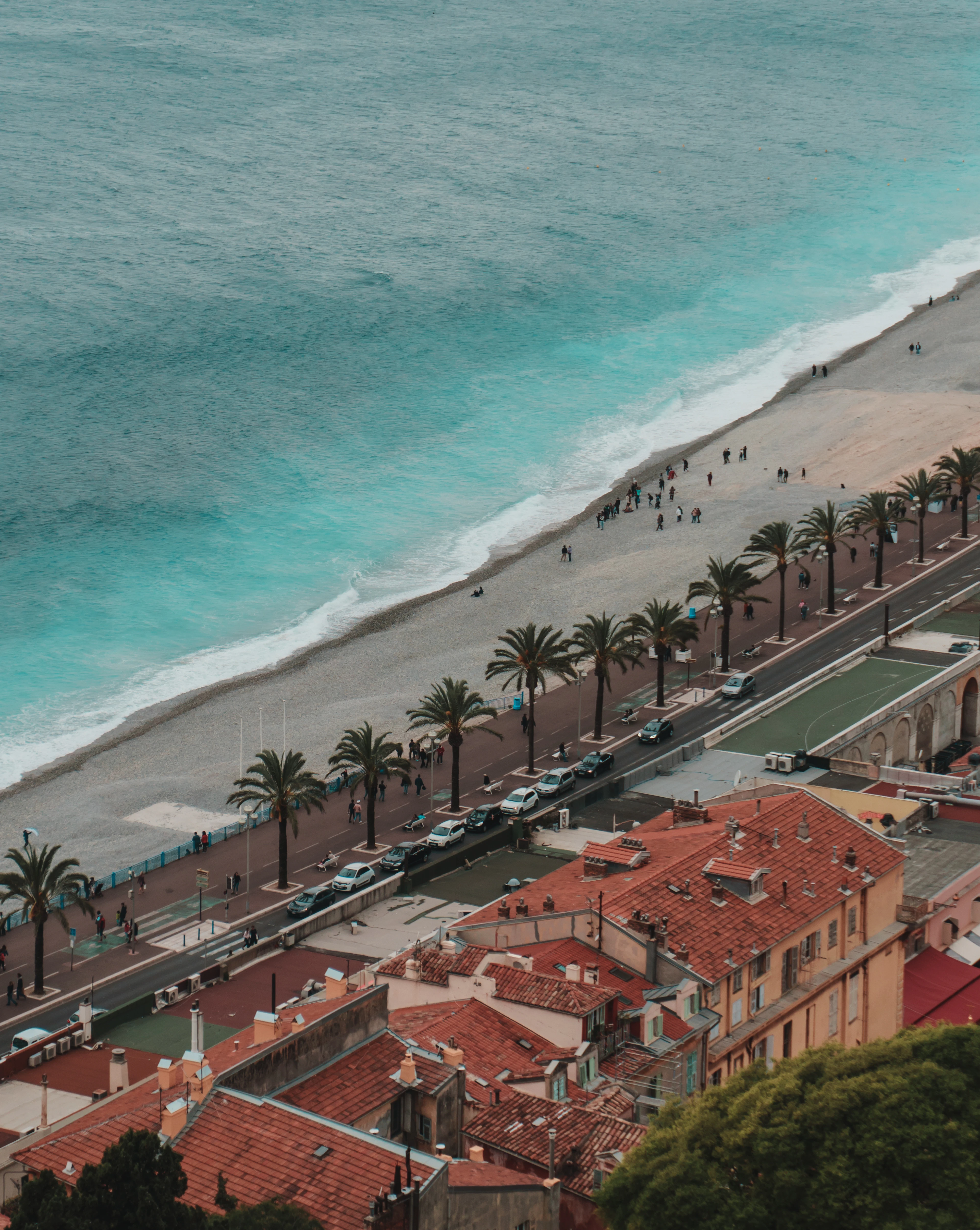 Aerial view of a curving bay lined with red-tiled rooftops, palm trees and a beach promenade beside turquoise water.