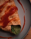 A close-up of a savory crepe or wrap featuring a soft, golden-brown exterior with a visible portion of green leafy vegetables peeking out. It is drizzled with a generous amount of reddish sauce, creating a visually appealing texture and color contrast. The scene is set on a simple, gray plate on a wooden surface.