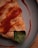 A close-up of a savory crepe or wrap featuring a soft, golden-brown exterior with a visible portion of green leafy vegetables peeking out. It is drizzled with a generous amount of reddish sauce, creating a visually appealing texture and color contrast. The scene is set on a simple, gray plate on a wooden surface.