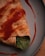 A close-up of a savory crepe or wrap featuring a soft, golden-brown exterior with a visible portion of green leafy vegetables peeking out. It is drizzled with a generous amount of reddish sauce, creating a visually appealing texture and color contrast. The scene is set on a simple, gray plate on a wooden surface.