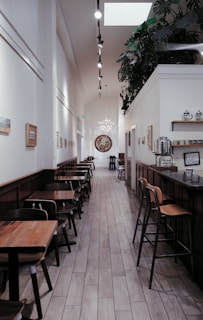 A long, narrow corridor of a modern café with wooden tables and chairs aligned along the walls. Potted plants adorn the top of a white partition. Soft lighting from hanging fixtures and spotlights creates a cozy atmosphere. Artwork and framed pictures decorate the walls.