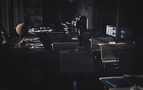 A dimly lit classroom with rows of old wooden desks and chairs. Piles of books and papers are scattered on the desks. A globe is placed on one of the front desks, and the classroom appears to be from an older era. Light is streaming in from the windows on the left, casting shadows across the room. At the back, there is a wooden dresser with a few drawers open and more books.