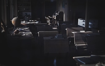 A dimly lit classroom with rows of old wooden desks and chairs. Piles of books and papers are scattered on the desks. A globe is placed on one of the front desks, and the classroom appears to be from an older era. Light is streaming in from the windows on the left, casting shadows across the room. At the back, there is a wooden dresser with a few drawers open and more books.