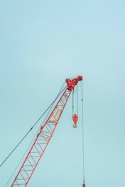 A red construction crane with a large hook hangs against a clear sky. The crane features a lattice boom design and is positioned at an angle with cables extending vertically.