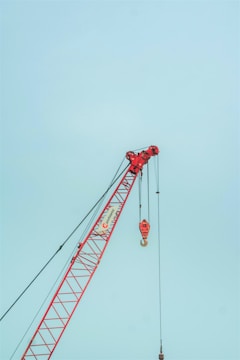 A red construction crane with a large hook hangs against a clear sky. The crane features a lattice boom design and is positioned at an angle with cables extending vertically.