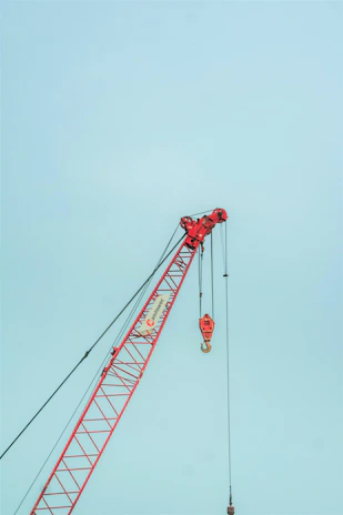 A side shot of a red truck crane extending its boom against a backdrop of steel beams.