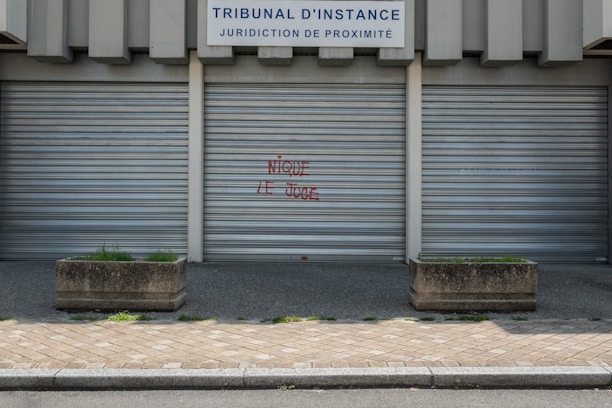 Metal shutters cover the entrance of a building labeled 'TRIBUNAL D'INSTANCE JURIDICTION DE PROXIMITÉ'. Red graffiti with the phrase 'NIQUE LE JUGE' is sprayed on one of the shutters. Two concrete planters with sparse greenery are placed on either side. The ground is paved with cobblestones.