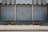Metal shutters cover the entrance of a building labeled 'TRIBUNAL D'INSTANCE JURIDICTION DE PROXIMITÉ'. Red graffiti with the phrase 'NIQUE LE JUGE' is sprayed on one of the shutters. Two concrete planters with sparse greenery are placed on either side. The ground is paved with cobblestones.