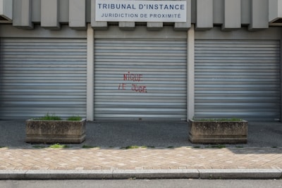 Metal shutters cover the entrance of a building labeled 'TRIBUNAL D'INSTANCE JURIDICTION DE PROXIMITÉ'. Red graffiti with the phrase 'NIQUE LE JUGE' is sprayed on one of the shutters. Two concrete planters with sparse greenery are placed on either side. The ground is paved with cobblestones.