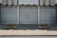Metal shutters cover the entrance of a building labeled 'TRIBUNAL D'INSTANCE JURIDICTION DE PROXIMITÉ'. Red graffiti with the phrase 'NIQUE LE JUGE' is sprayed on one of the shutters. Two concrete planters with sparse greenery are placed on either side. The ground is paved with cobblestones.
