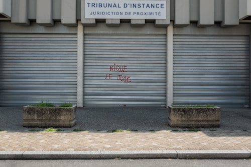 Metal shutters cover the entrance of a building labeled 'TRIBUNAL D'INSTANCE JURIDICTION DE PROXIMIT&Eacute;'. Red graffiti with the phrase 'NIQUE LE JUGE' is sprayed on one of the shutters. Two concrete planters with sparse greenery are placed on either side. The ground is paved with cobblestones.