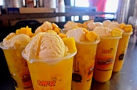 Multiple cups filled with a yellow beverage, topped with scoops of creamy ice cream and chunks of mango. The cups have a vibrant design with the text 'MFR' and other writings, placed on a reflective surface with a blurred background.