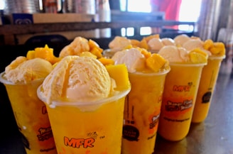 Multiple cups filled with a yellow beverage, topped with scoops of creamy ice cream and chunks of mango. The cups have a vibrant design with the text 'MFR' and other writings, placed on a reflective surface with a blurred background.