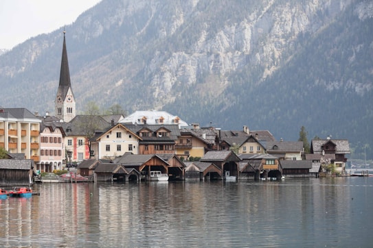 A picturesque lakeside village with traditional alpine architecture nestled against a backdrop of misty mountains. The buildings feature steeply pitched roofs and wooden facades, with a tall church spire rising prominently. The calm lake reflects the charming structures and creates a serene atmosphere.