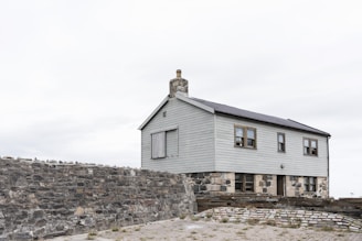 A simple two-story house with light gray wooden paneling sits atop a solid stone foundation. There is a chimney on the roof, and several small windows are visible. The building is enclosed by a rough stone wall, and the foreground shows a paved area with sparse grass.