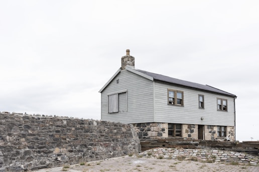 A simple two-story house with light gray wooden paneling sits atop a solid stone foundation. There is a chimney on the roof, and several small windows are visible. The building is enclosed by a rough stone wall, and the foreground shows a paved area with sparse grass.