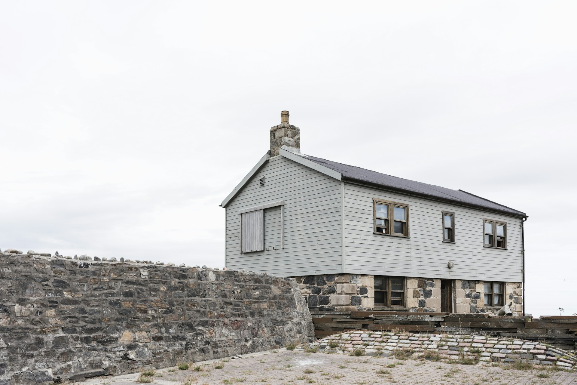 A simple two-story house with light gray wooden paneling sits atop a solid stone foundation. There is a chimney on the roof, and several small windows are visible. The building is enclosed by a rough stone wall, and the foreground shows a paved area with sparse grass.