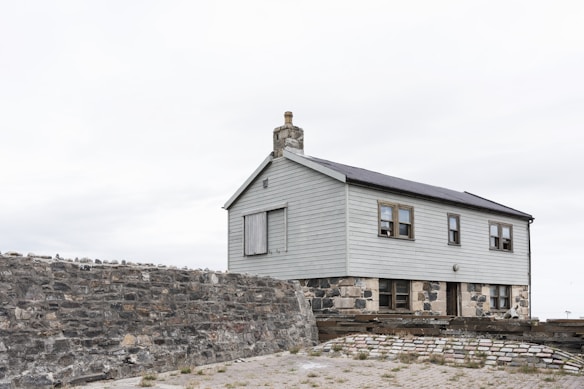 A simple two-story house with light gray wooden paneling sits atop a solid stone foundation. There is a chimney on the roof, and several small windows are visible. The building is enclosed by a rough stone wall, and the foreground shows a paved area with sparse grass.