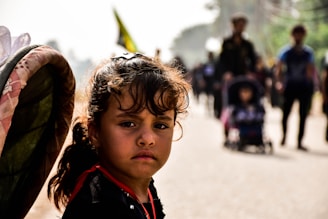 A young girl with curly hair looks directly at the camera with a serious expression. She stands outdoors among a group of people, some of whom are out of focus in the background. The light creates a soft glow around her face, highlighting her features.